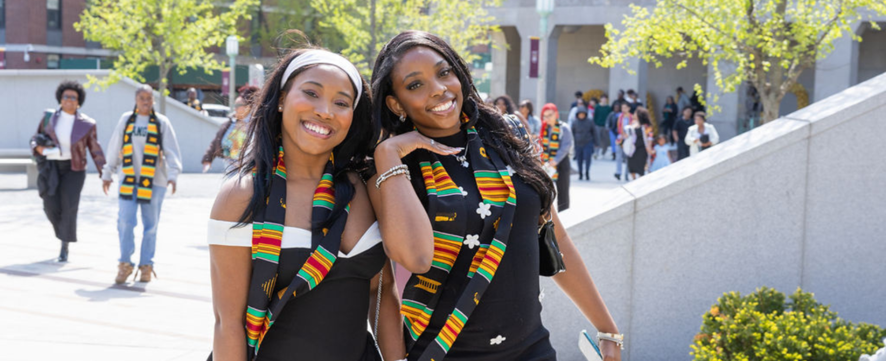 Two women in black dresses with colorful stoles, smiling outdoors.